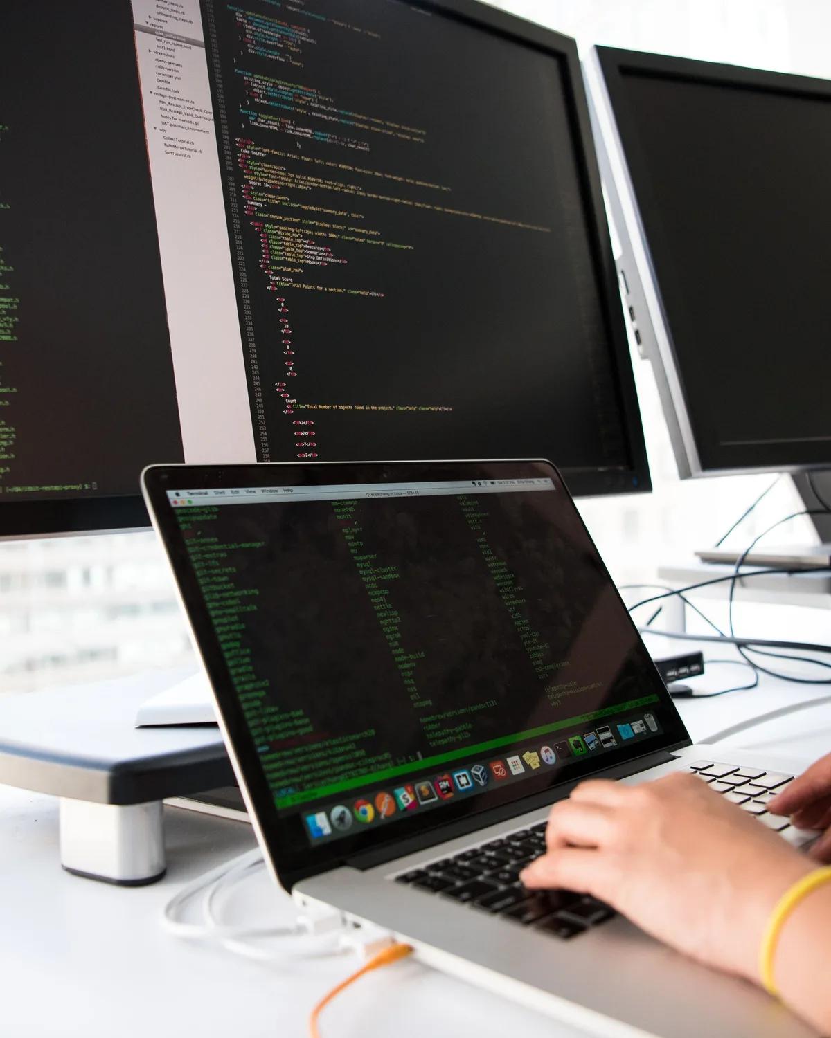 Code on a laptop and two monitors, with a hand on the keyboard reviewing it.
