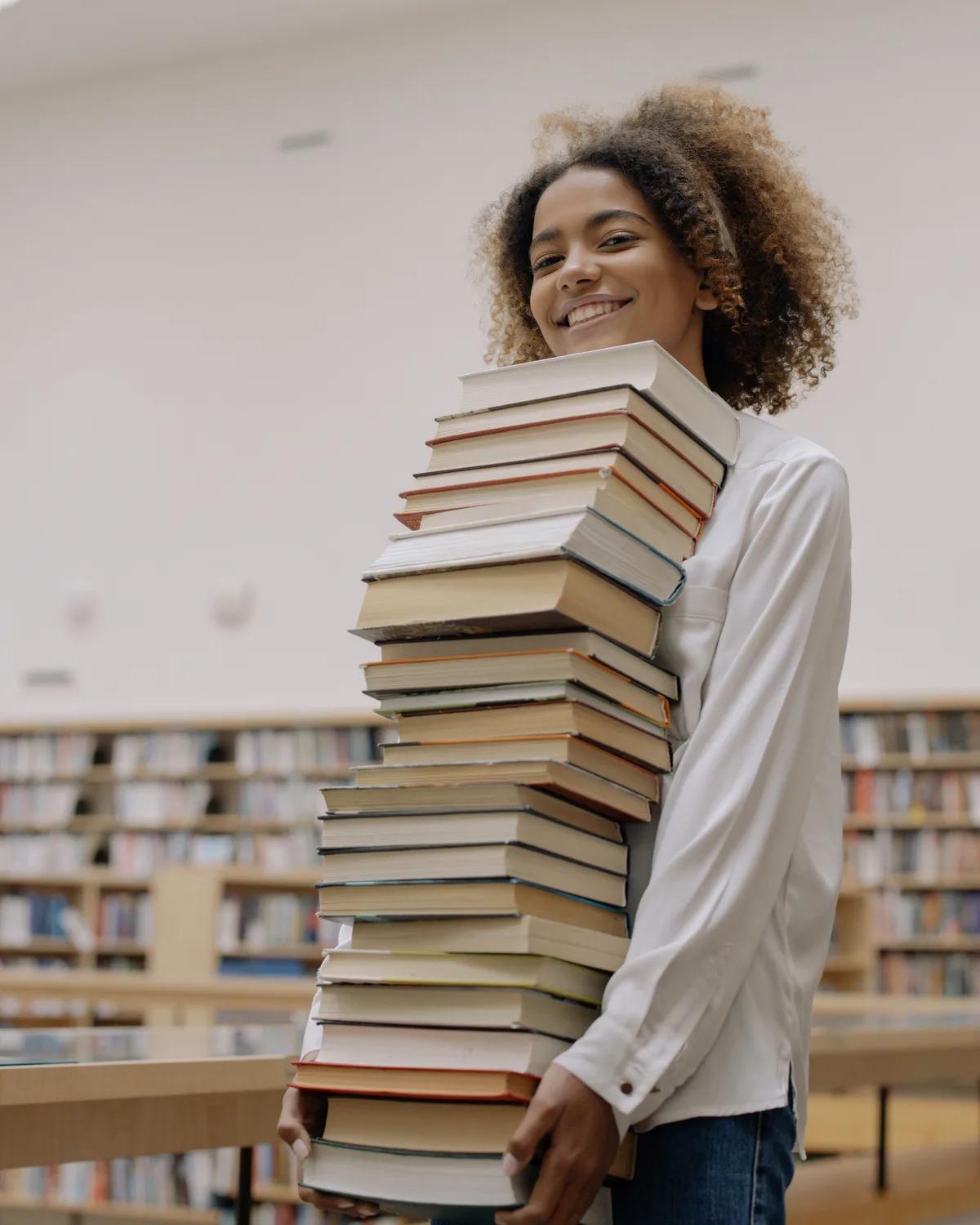 Someone carrying a tall stack of reference books through a library, smiling.