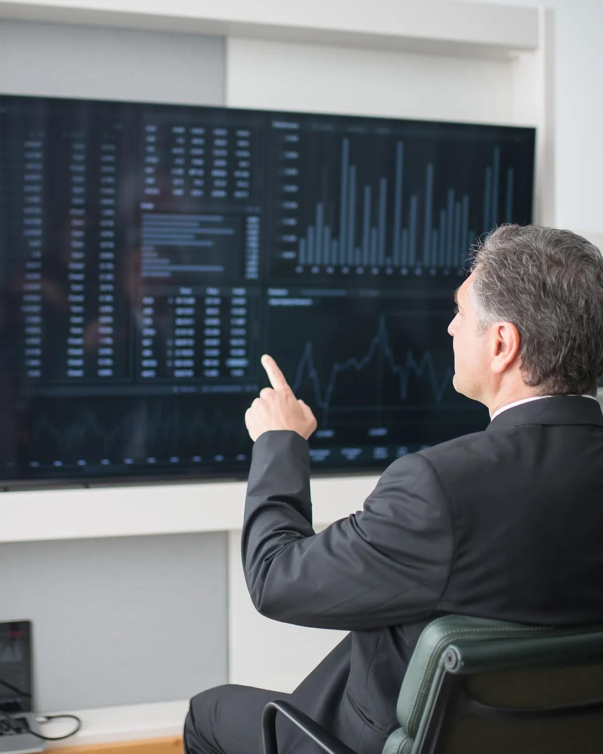 A manager pointing at a wall screen of live business dashboards in an office.